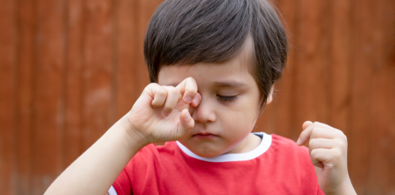 A young boy in a red and white top rubbing his eyes outside.