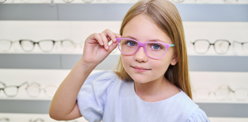 A young girl trying on a pair of glasses while looking at the camera while in an opticians practice.